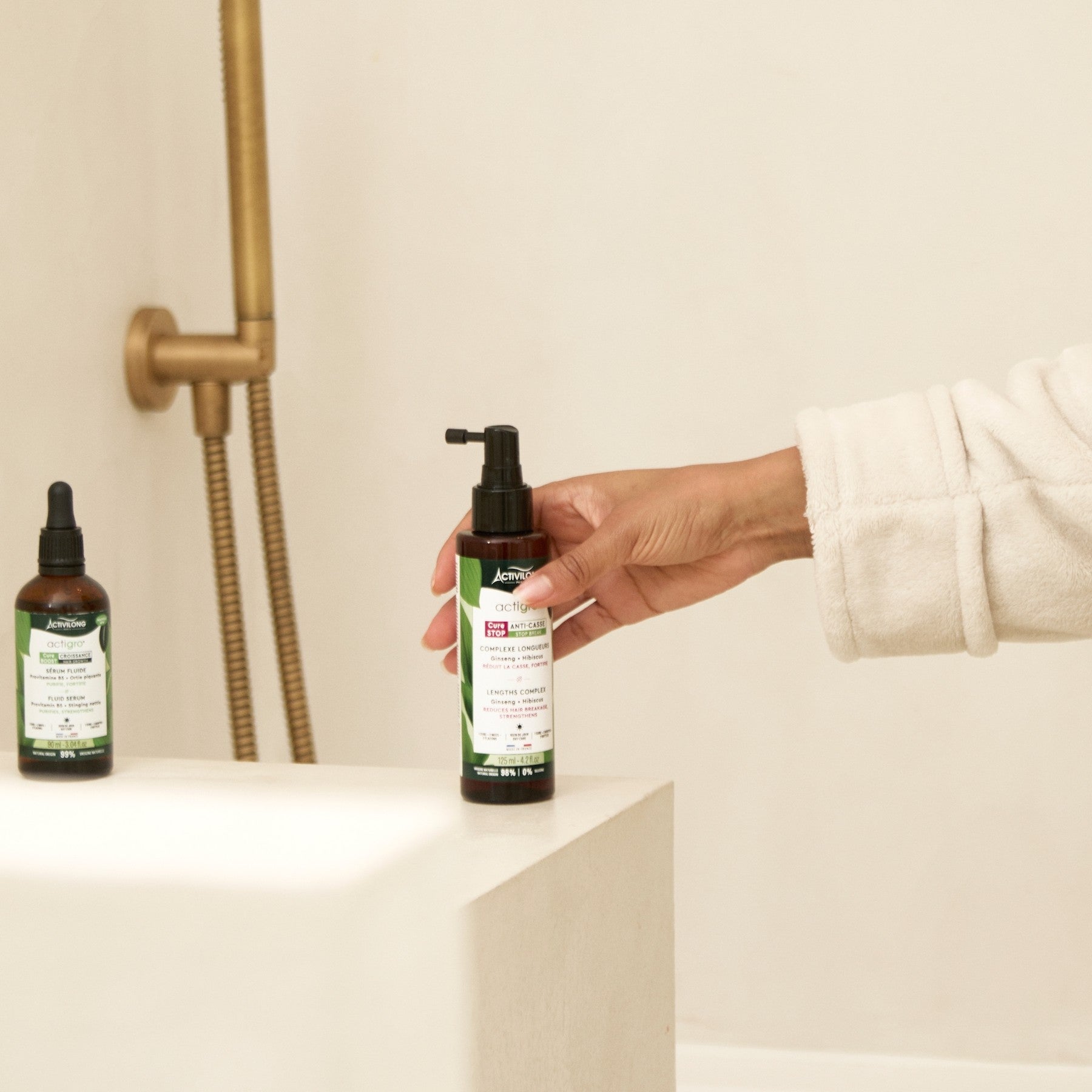 Hand holding an ACTIVILONG ACTIGRO Sérum Complexe Longueurs 125ml bottle by Afro Beauty Store with another bottle on a shelf in a bathroom setting
