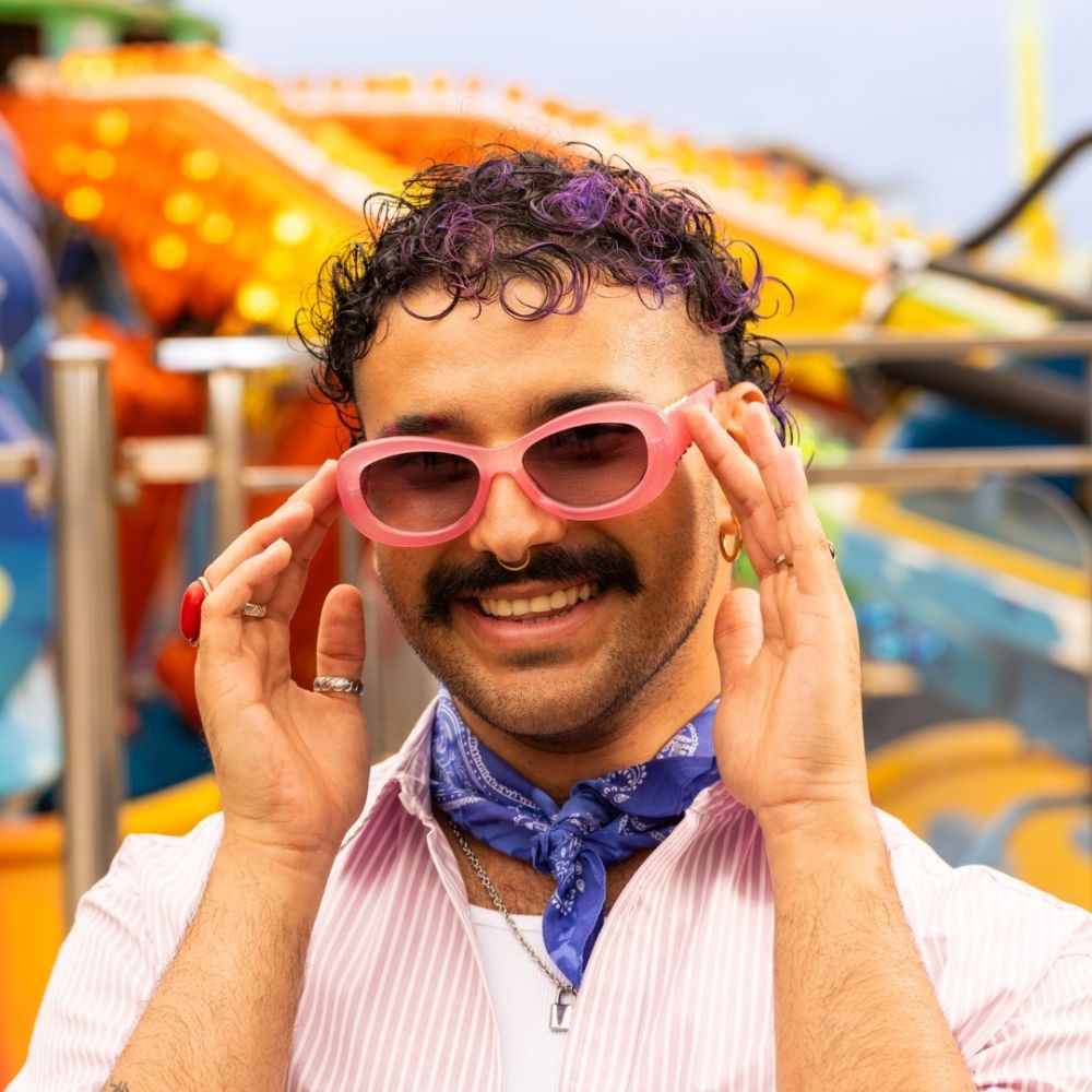 Crazy Color's person wearing pink sunglasses and a blue bandana, with colorful fairground rides in the background by Afro Beauty Store