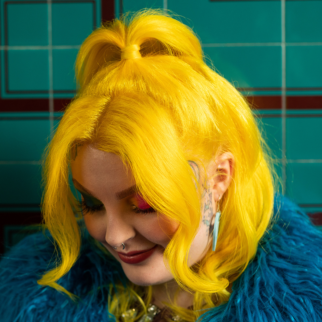 Crazy Color's Person with bright yellow hair styled in a high bun, wearing a blue fur coat against a tiled wall by Afro Beauty Store
