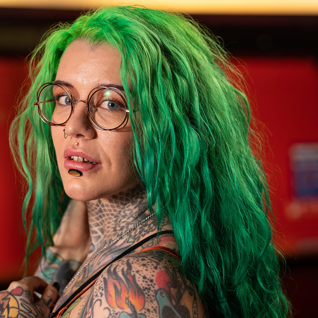 Crazy Color's Person with green hair and tattoos wearing glasses, against a blurred red background by Afro Beauty Store