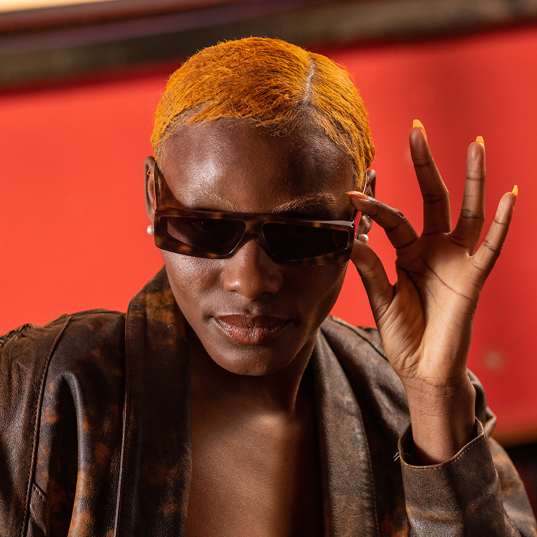 Crazy Color's Person with orange hair wearing sunglasses and a brown jacket against a red background by Afro Beauty Store
