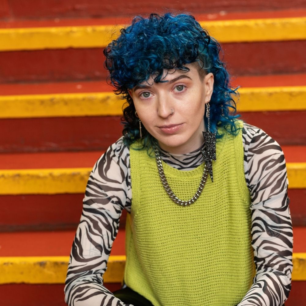 Crazy Color's Person with blue hair and a green sweater sitting on colorful steps by Afro Beauty Store
