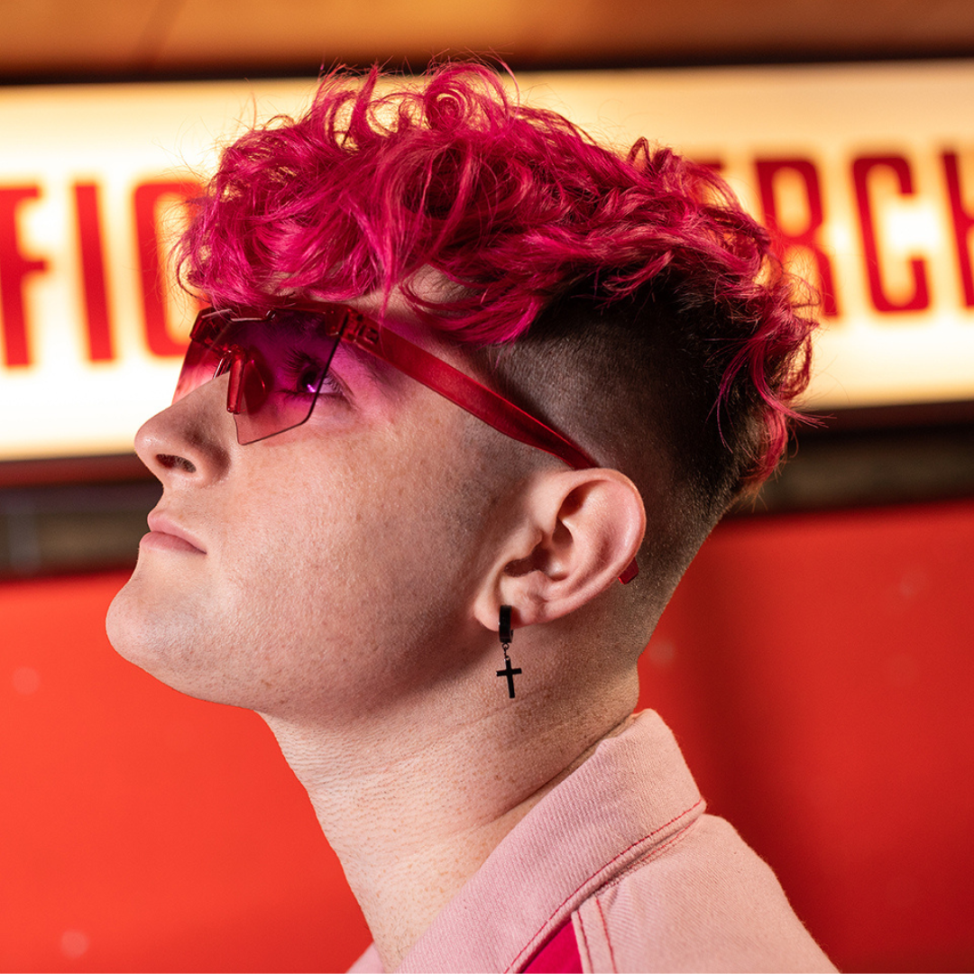 Crazy Color's Person with pink hair and sunglasses looking to the side, against a red and yellow background by Afro Beauty Store