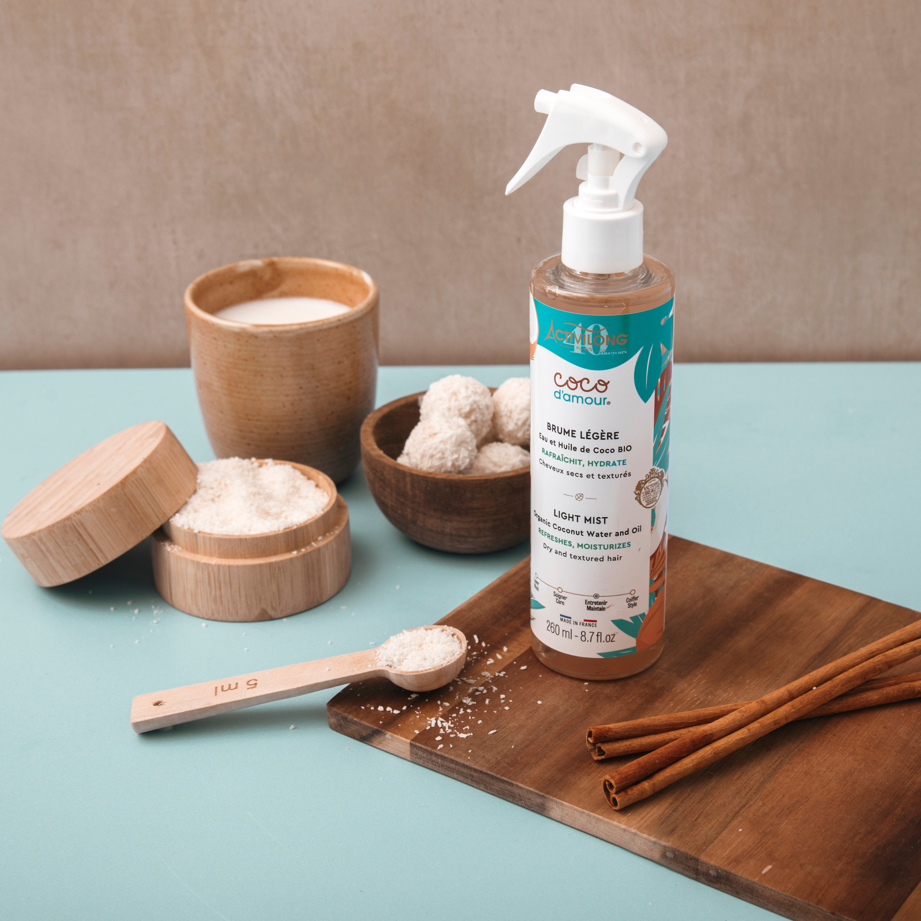 Bottle of ACTIVILONG COCO D’AMOUR Brume Légère 260ml by ABS France Wholesale on a wooden board with wooden containers and cinnamon sticks on a teal surface.