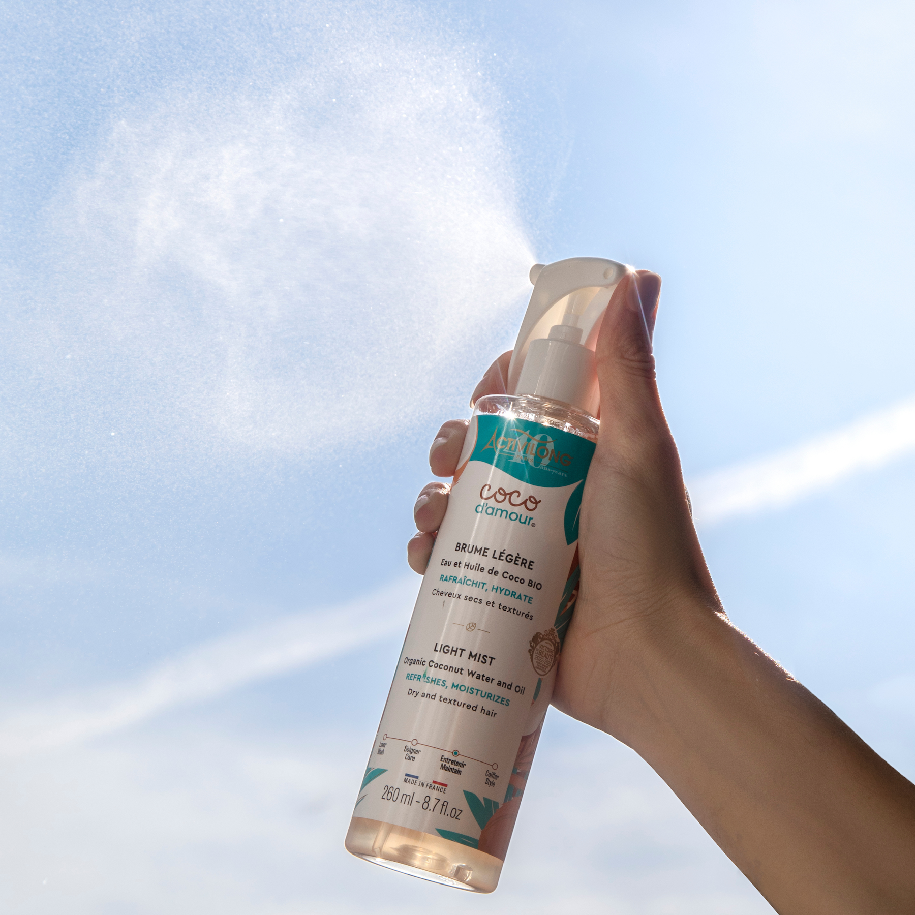 Hand holding a bottle of ACTIVILONG COCO D’AMOUR Brume Légère 260ml by ABS France Wholesale against a blue sky background
