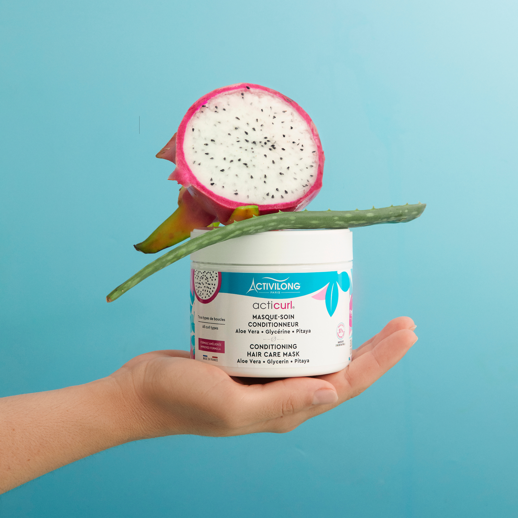 Hand holding a ACTIVILONG ACTICURL Masque 300ml jar by Afro Beauty Store with a dragon fruit on a blue background
