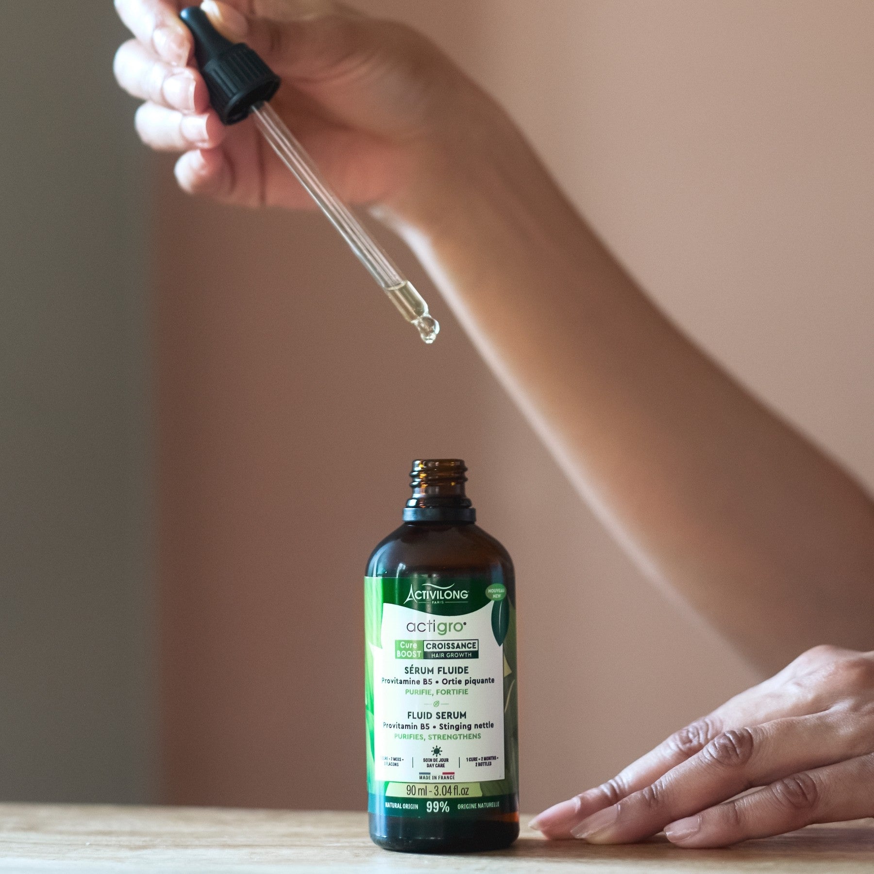Hand holding a dropper over a Bottle of ACTIVILONG ACTIGRO Sérum Fluide 90ml by Afro Beauty Store on a wooden surface.